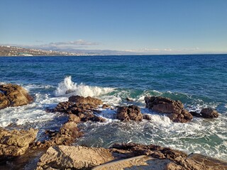 Waves crashing on sunlit, jagged rocks on the coast of Opatija, Croatia, with the deep blue Adriatic Sea stretching towards the city in the distance.