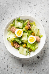 Fresh salad with tuna, cucumber ribbons, boiled eggs, olives, and lettuce in a white bowl on a gray surface. Healthy protein-rich meal, top view, vibrant food styling