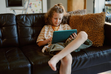 A young boy sits comfortably on a black couch, absorbed in his tablet. He is relaxed, wearing...