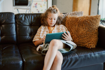 A young boy sits on a comfortable couch with a decorative pillow, focused on his tablet. He wears casual clothes and is engaged in his activity, creating a peaceful atmosphere in the living room.