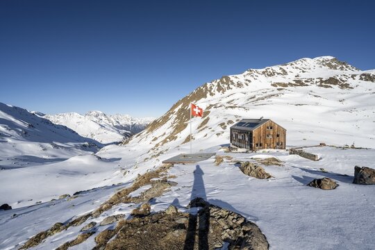 Swiss flag and mountain hut Keschh&uuml;tte in front of mountain landscape in winter, behind summit of the Berg&uuml;ner St&ouml;cke Tinzenhorn and Piz Ela, B&uuml;ndner Haute Route, Albula Alps, Rhaetian Alps, Grisons, Eastern Switzerland, Switzerland