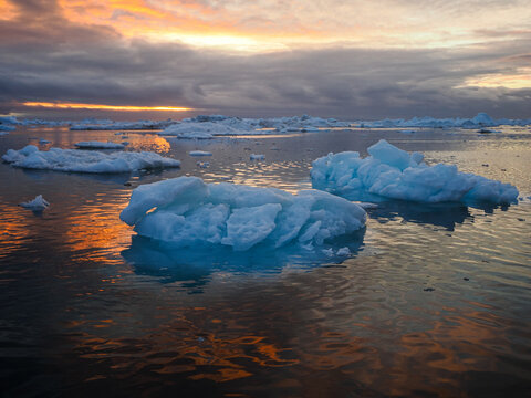 Floating icebergs in calm Arctic water reflecting vibrant sunset sky and dramatic clouds