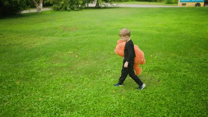 Young boy walking slowly across open grassy field while holding large orange teddy bear, looking downward thoughtfully with yellow bus partially visible in distant background near road