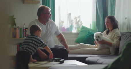Father and boy interacting with toy train on coffee table, mother holding white dog on sofa in background, intimate family connection and bonding indoors