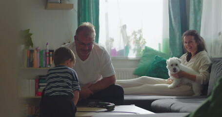 Father and boy interacting with toy train on coffee table, mother holding white dog on sofa in background, intimate family connection and bonding indoors