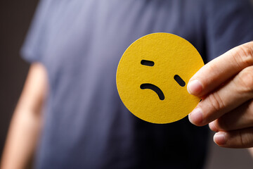 A hand holds a yellow circle with a disappointed facial expression, conveying feelings of sadness,...