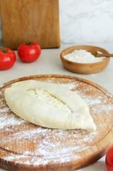 Raw khachapuri on a wooden cutting board with red tomatoes in the kitchen. The process of making bread with cheese. The concept of traditional Georgian food. Vertical orientation. Selective focus.