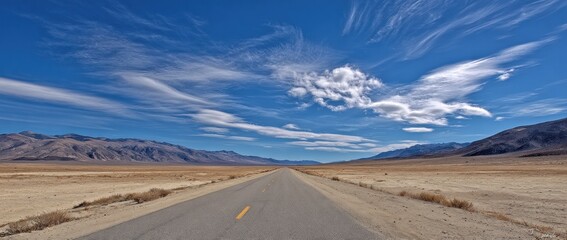 A long straight road stretching through a dry landscape with distant mountains and a bright blue sky filled with wispy clouds
