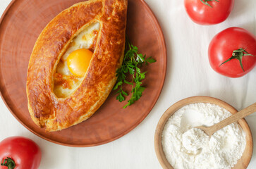 Adjarian khachapuri with egg yolk on a ceramic plate with tomatoes on the table. The concept of traditional Georgian pastries. Horizontal orientation. Top view.