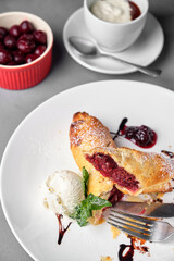 Close-up shot of a delicious cherry strudel, served with a scoop of ice cream and mint leaves, on a white plate.
