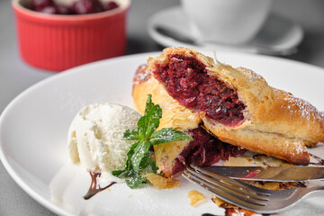 Close-up shot of a delicious cherry strudel, served with a scoop of ice cream and mint leaves, on a white plate.
