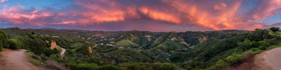 Panorama view lush hills at dusk under a radiant, pink-orange sky. Trails meander through the landscape. Houses dot the distant terrain
