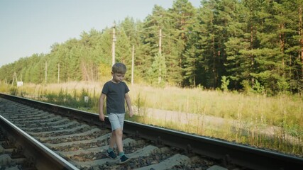 young boy wearing grey shorts and dark shirt walking carefully on gravel-covered rail track with lush forest and utility poles in background under clear blue sky and bright sunlight