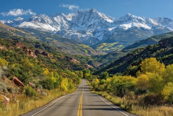 Fototapeta premium Scenic highway stretching into mountains with autumn colors, and snowy peaks against blue sky
