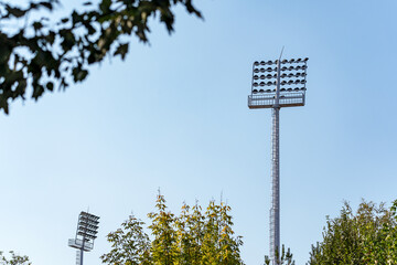 A low-angle shot captures two tall stadium floodlight towers reaching towards a clear, bright blue sky, in the foreground green trees. 