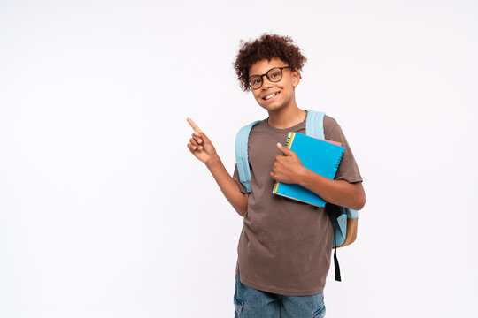 Smiling African-American preteen boy male elementary primary school pupil holding copybooks with backpack showing pointing index finger at copy space for promotional ad on white background.