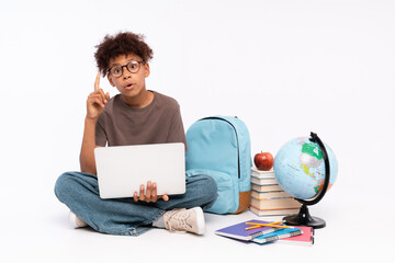 Amazed African-American school boy elementary primary pupil sitting near books copybooks backpack globe holding using laptop for e-learning pointing index finger up isolated on white background.