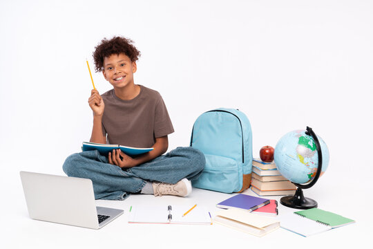 Smiling African-American school boy elementary primary pupil surrounded copybooks books backpack globe laptop holding reading book having an idea pointing index finger up isolated white background