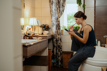 A young woman sits on the edge of a bathtub in a well-lit bathroom, brushing her teeth while checking her phone. The space is decorated with plants and personal items.