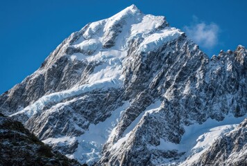 Naklejka premium A sharp, snow-covered mountain peak under a bright blue sky, revealing rock faces and glacial ice flow with a hint of vegetation at the bottom