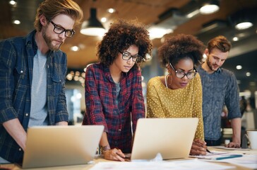 Four young adults collaborate over laptops and papers in a bright, open office setting, discussing and working together as a team