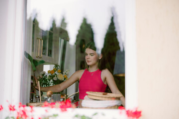 A young woman in a bright pink top prepares baked goods in a kitchen filled with natural light. Fresh flowers and culinary items decorate the warm atmosphere.