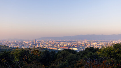 Fototapeta premium Cityscape of Kyoto seen from Yotsutsuji Intersection trail on Mount Inari, with foreground trees, layered urban skyline and distant mountains at sunrise