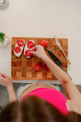 A person is skillfully assembling a sandwich, placing fresh tomato slices on bread at a kitchen table. The bright atmosphere enhances the culinary activity.