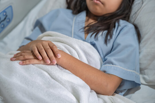 Close-up of a young girl lying in a hospital bed with her hands folded on a white blanket, symbolizing illness, recovery, or patient care in a healthcare or medical environment. - Powered by Adobe
