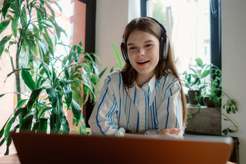 In a sunlit room filled with indoor plants, a young girl engages happily in an online class while wearing headphones and smiling at her laptop, showcasing the joy of learning.
