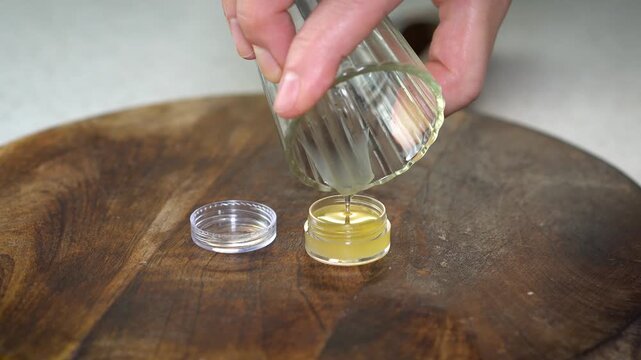 Pouring natural DIY lip balm from glass jar into small transparent container on dark wooden background. Organic skincare, handmade cosmetics, eco-friendly beauty, and artisan style.