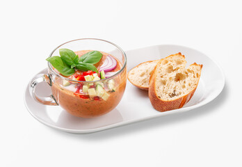 Traditional Spanish gazpacho cold tomato soup served in a glass cup with fresh basil, diced vegetables, red onion and crusty baguette slices on a white plate, isolated on white background