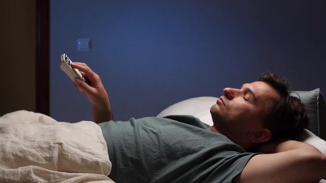 Man watching tv lying in bed at night using remote control