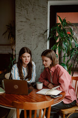 Inside a cozy cafe, two young women are focused on a laptop while sharing ideas. A warm atmosphere offers a perfect backdrop for their study session and brainstorming activities.
