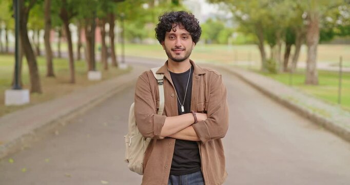 Indian happy young adult gen z man male look camera cross arm standing outdoor area. Smart cool proud pupil boy guy wear bag fold hand staring cam do casual pose enjoy new day outside place