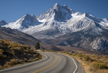 Naklejka premium A curving asphalt road leads to a snowy mountain range beneath a clear blue sky, showcasing a scenic landscape view