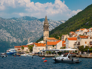 Sailing boats and waterside stone buildings on the Bay of Kotor shoreline at Perast in Montenegro. Rugged mountains in the background. Clear blue sky.