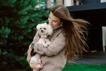 A young woman with long hair smiles as she cuddles a small dog outdoors. The background includes lush green trees and modern architecture, creating a joyful atmosphere.