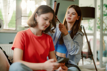 Two friends enjoy a relaxed afternoon in a cozy room. One styles the other's hair while she checks her phone, creating a fun and casual atmosphere.