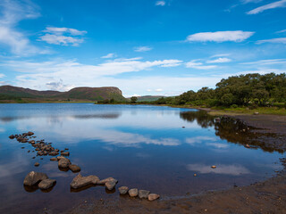 The calm, tranquil waters of a Loch Brora shoreline against rolling rugged hills and heather-clad moorlands of the Highlands in Northern Scotland. Bright blue sky.