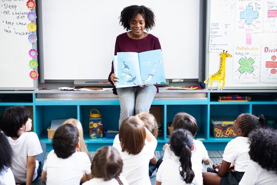 Teacher reading to diverse group of children. Classroom setting with teacher and students. Engaged children listening to teacher reading a book. Black teacher teaching nursery school class of children - Powered by Adobe