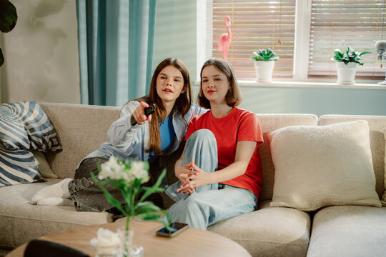 Two young women sit comfortably on a couch in a cozy living room, focused on a movie. One points at the screen, while the other relaxes, creating an inviting atmosphere.
