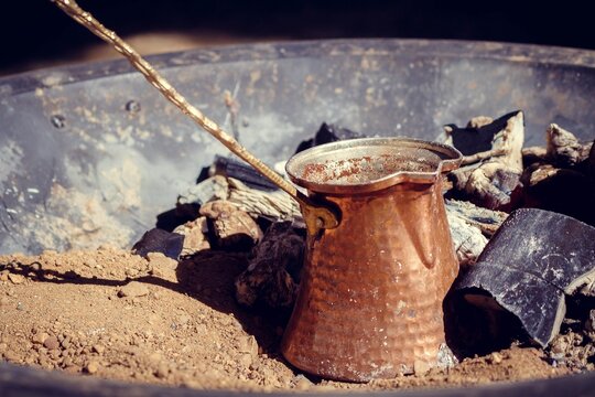 A traditional copper cezve with long handle brewing strong arabic coffee on hot embers in a metal tray