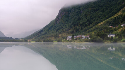 Serene waters of Bergheimsvatnet lake mirror the lush green mountains and quaint village in Norway. Morning fog enhances the peaceful atmosphere, creating a picturesque natural landscape.