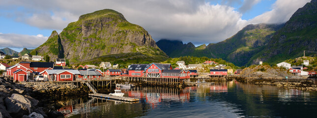 Vibrant fishing cabins reflect in the calm waters of A Lofoten, Norway. Majestic mountains rise in the background under a partly cloudy sky, creating a breathtaking coastal landscape during daylight.