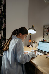 A young student focuses on her studies at a wooden desk during nighttime. She writes in a notebook while a laptop illuminates the space with a warm glow.