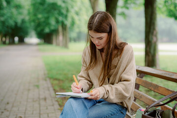 Obraz premium A young woman sits on a wooden bench in a scenic park, focused on writing in her notebook. Lush green trees surround her, creating a serene atmosphere.