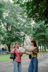 Fototapeta premium Two friends stand in a green park, engaging in a lively conversation while one hands over a bag of snacks. The setting features lush trees and a nearby amusement ride.