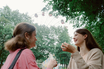 Two young women share a joyful moment at an amusement park, laughing and holding snacks. A ferris wheel looms in the background, enhancing the lively atmosphere of the day.