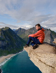 A hiker sits on the edge of a rocky precipice on the Ryten Kvalvika Beach Trail, admiring breathtaking views of the turquoise waters and rugged landscape of Lofoten, Norway.